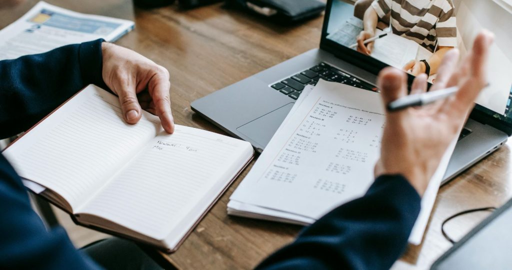 Fotografía de las manos de una persona con un cuaderno frente a un computador donde se ve un estudiante