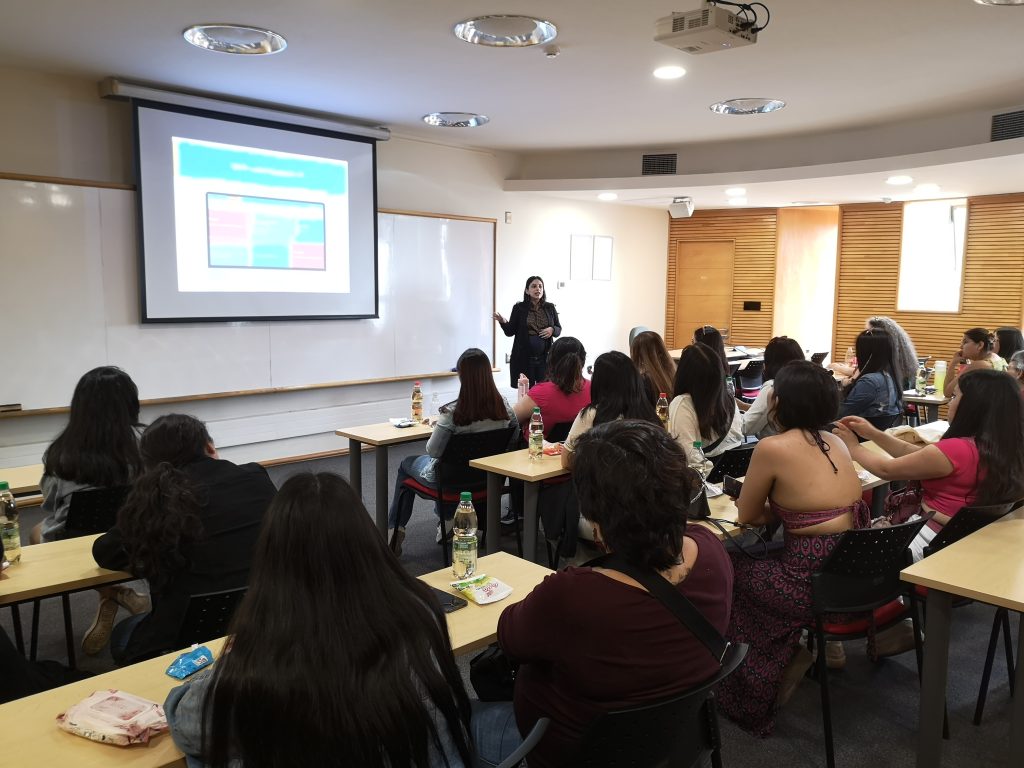 Fotografía de una sala de clases, se ven decenas de personas sentadas y una persona en frente proyectando una presentación