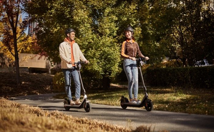 Fotografía de dos personas en un scooter cada una, andando en un camino de cemento alrededor de una zona de pasto y árboles